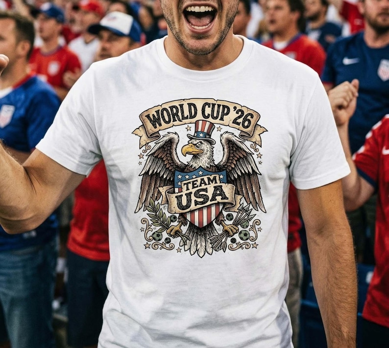 May include: White t-shirt with a graphic featuring an eagle wearing a top hat, wings spread, and a shield with the text "TEAM USA." The shirt also has the text "WORLD CUP '26" in a banner above the eagle.