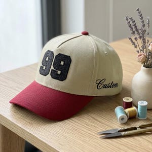 Puede incluir: Gorra de béisbol beige con visera burdeos y botón rojo. El número "99" está bordado en negro en la parte delantera, con la palabra "Custom" en cursiva. La gorra está sobre una superficie de madera.