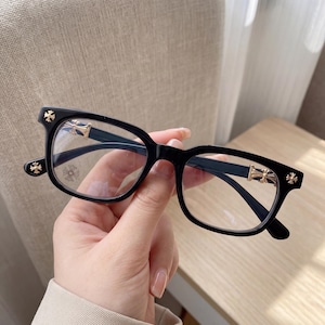 May include: Black eyeglasses with a rectangular frame. The temples and hinges feature gold-colored cross and floral designs. The lenses are clear, and the glasses are held by a hand.