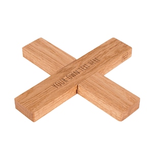 Pode incluir: Um descanso de panela de madeira castanho-claro em forma de X, construído a partir de duas barras retangulares que se cruzam. A superfície apresenta o texto gravado "YOUR OWN TEXT HERE" e um pequeno logótipo quadrado com "oru nics".