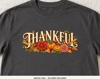 Könnte beinhalten: Ein dunkelgraues T-Shirt mit dem Wort THANKFUL in cremefarbener Schrift. Der Text ist mit einer herbstlichen Grafik verziert, die einen orangefarbenen Kürbis, rote und gelbe Dahlien, grüne Blätter und goldene Weizenähren zeigt.