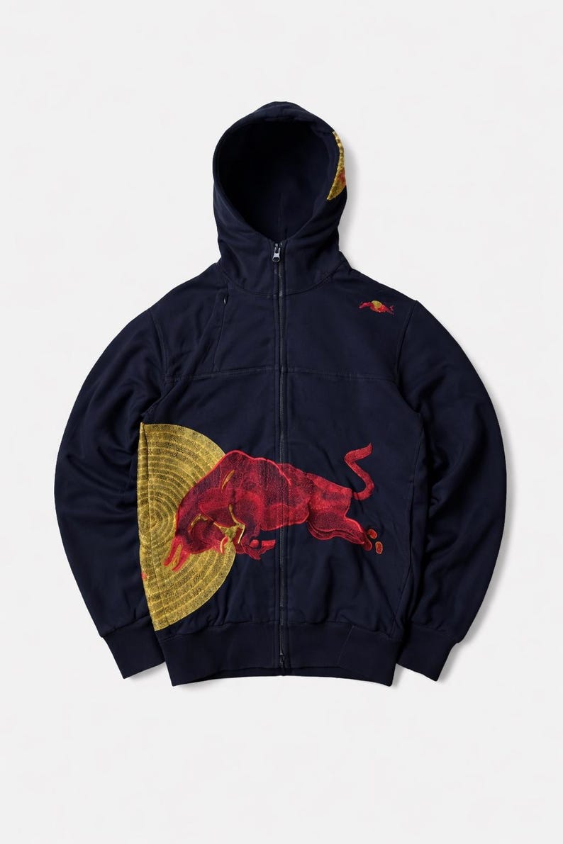 Puede incluir: Sudadera con capucha azul marino con cremallera y un gran gr&aacute;fico de toro rojo en la parte delantera, sobre un dise&ntilde;o circular dorado. La sudadera tiene una capucha con forro dorado y un peque&ntilde;o logotipo de toro rojo en el pecho superior derecho.