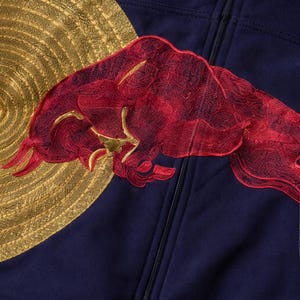 Puede incluir: Sudadera con capucha azul marino con cremallera, adornada con un sol bordado en oro y un logotipo de toro bordado en rojo. La sudadera tiene una cremallera completa y una tela suave.
