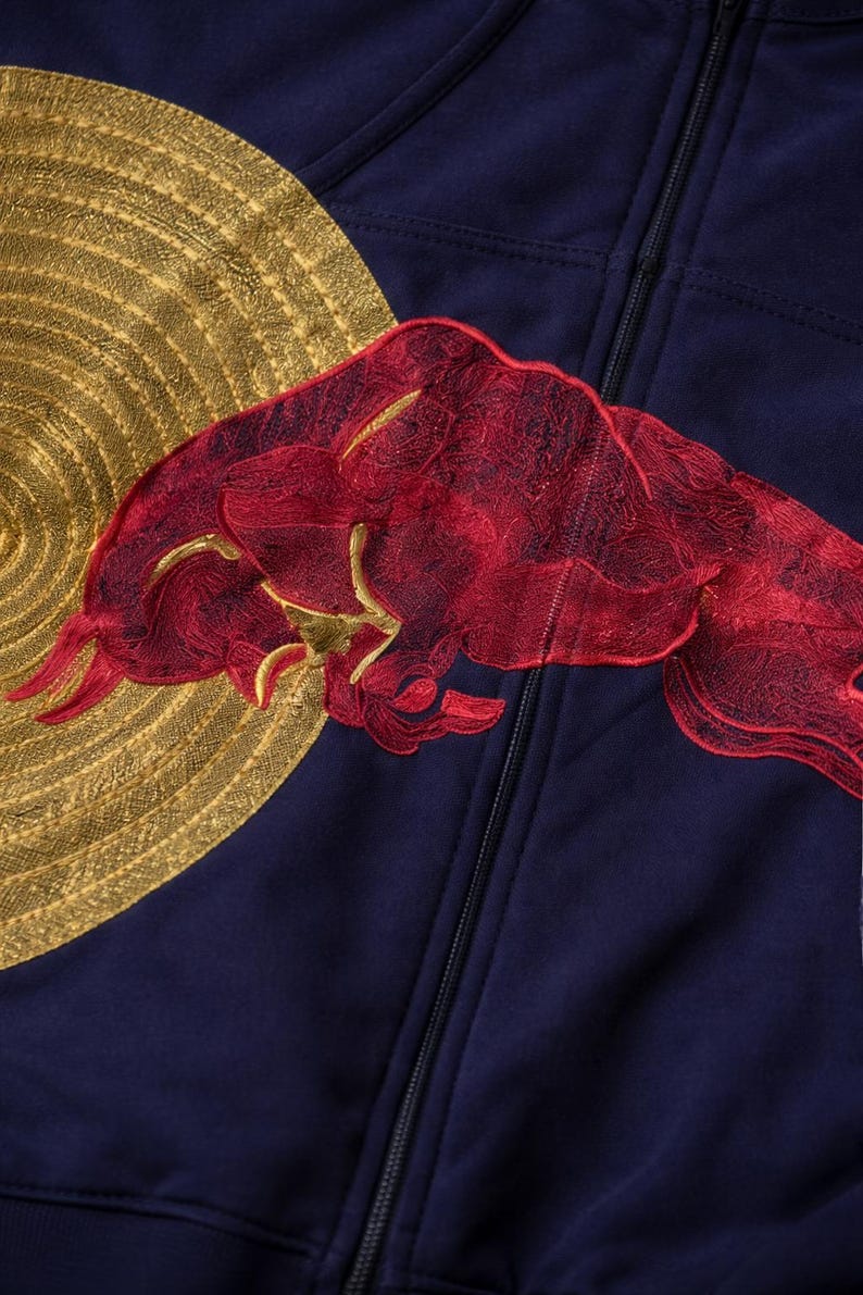 Puede incluir: Sudadera con capucha azul marino con cremallera, adornada con un sol bordado en oro y un logotipo de toro bordado en rojo. La sudadera tiene una cremallera completa y una tela suave.
