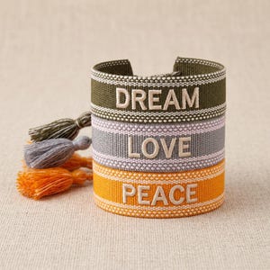 Könnte beinhalten: Gestapelte Stoffarmbänder in Olivgrün, Grau und Orange, jeweils mit einem Wort bestickt: DREAM, LOVE und PEACE. Die Armbänder haben weiße Nähte und werden von passenden Quastenanhängern begleitet.