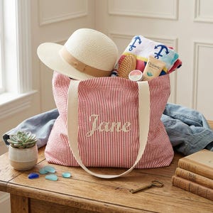 Könnte beinhalten: Eine rot-weiß gestreifte Tragetasche mit dem gestickten Namen "Jane". Die Tasche enthält einen Strohhut, ein Strandtuch, eine Haarbürste und Hautpflegeprodukte. Daneben steht ein kleiner Topf mit einer Sukkulente.