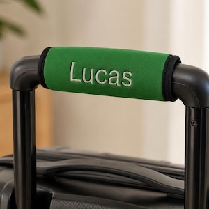 Op de afbeelding: Een groene bagagehandgreep met de naam "Lucas" in het wit geborduurd. De handgreep zit op een zwarte koffer. De handgreep is ontworpen voor comfort en personalisatie.