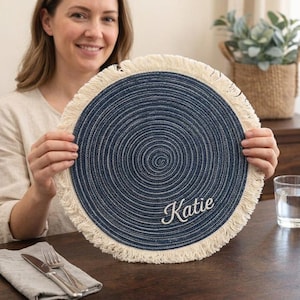 Könnte beinhalten: Ein runder, marineblauer Platzdeckchen mit Spiralmuster und cremefarbenem Fransen. Der Name "Katie" ist in weißer Schrift gestickt. Eine Person hält den Platzdeckchen, und ein Tischgedeck ist sichtbar.