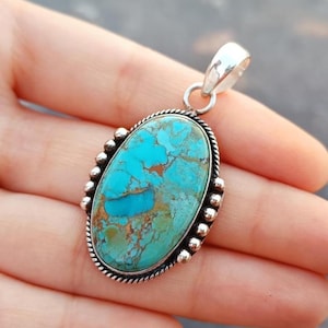 Könnte beinhalten: Ovaler Türkis-Anhänger mit silberner Öse und dekorativem Silberrand. Der Türkisstein hat eine gesprenkelte blau-grüne Farbe mit braunen und goldenen Adern. Der Anhänger wird in einer Hand gehalten.