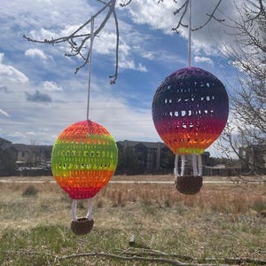May include: Two crocheted hot air balloons hanging from a tree branch against a blue sky with white clouds. The balloon on the left is green, yellow, orange, and red. The balloon on the right is purple, pink, and orange.