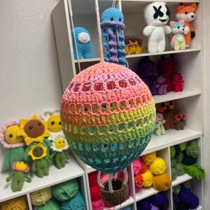 May include: A rainbow crocheted hot air balloon with a brown crocheted basket hanging from it. The balloon is hanging from the ceiling and there are yarn balls in the background.