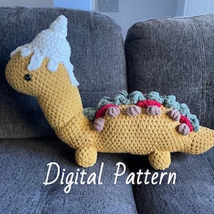Puede incluir: Juguete de peluche de dinosaurio de crochet con una cabeza blanca de crochet y un cuerpo amarillo, verde y rojo. El juguete está sentado en un sofá gris. El texto "Digital Pattern" es visible debajo del juguete.
