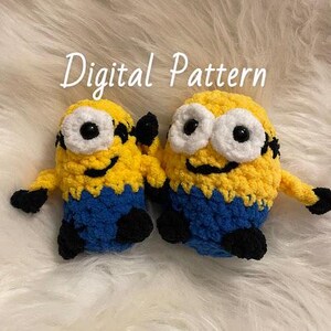 Puede incluir: Dos peluches de crochet con forma de los Minions, personajes de la película Gru, mi villano favorito. Los peluches son amarillos con monos azules y pelo negro. Tienen ojos blancos y pupilas negras. Los peluches están sentados sobre una superficie blanca y esponjosa.