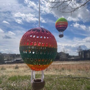 May include: Two crocheted hot air balloons hanging from a tree branch against a blue sky with white clouds. The larger balloon is a rainbow of colors, while the smaller balloon is green, yellow, and orange.
