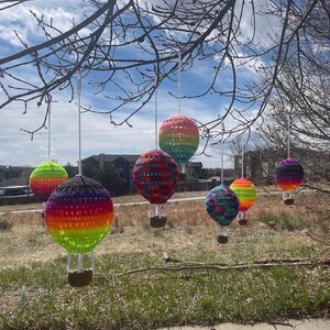 May include: Six colorful crocheted hot air balloons hanging from a tree branch against a blue sky with white clouds. The balloons are made of yarn in various colors, including red, orange, yellow, green, blue, purple, and black.