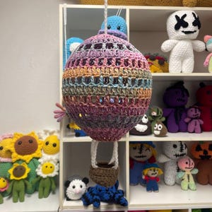 May include: A crocheted hot air balloon with a rainbow colored balloon and a brown basket. The balloon is hanging from the ceiling and there are other crocheted toys on shelves in the background.