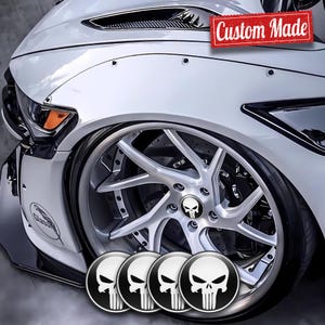 Könnte beinhalten: Ein weißes Auto mit individuellen silbernen Felgen mit einem Totenkopf-Logo. Das Bild zeigt eine Nahaufnahme des Vorderrads des Autos und einen Satz von vier Totenkopf-Logo-Emblemen. Der Text "Custom Made" ist auf einem roten Banner zu sehen.