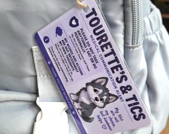 May include: A rectangular medical communication alert tag attached to a light grey backpack with twine. The white tag features purple text reading TOURETTE'S & TICS, an illustration of a grey puppy, and a message explaining involuntary tics.
