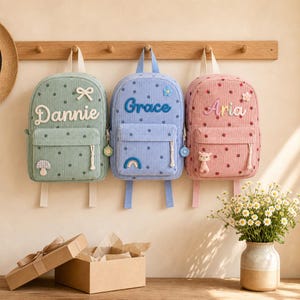 May include: Three corduroy backpacks in sage green, light blue, and dusty pink, each featuring a name embroidered in white or blue, small floral patterns, and unique decorative patches including a mushroom, rainbow, and small animal figure.