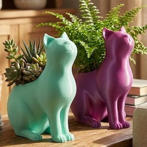 May include: Two cat-shaped planters in mint green and purple, each holding plants. The planters sit on a wooden table, with a cozy living room setting in the background. The cat planters are a unique home decor item.