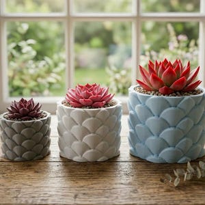 May include: A set of three ceramic mugs with a speckled, cream-colored glaze. Each mug features a different hand-painted design: a blue and green leaf, a yellow flower, and a red berry. The mugs have a simple, rounded shape and a comfortable handle.