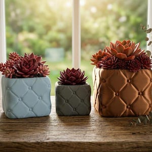 May include: Three square planters with succulents on a wooden surface. The planters are blue, grey, and bronze, each with a diamond-quilted pattern. The succulents are deep red, and the background is a blurred view of a window, with natural light.