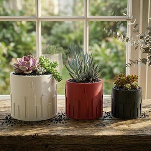 May include: Three cylindrical planters with succulents on a wooden surface. The planters are cream, red, and black, each with a cityscape design. The plants include a purple succulent, a green succulent, and a variety of succulents.