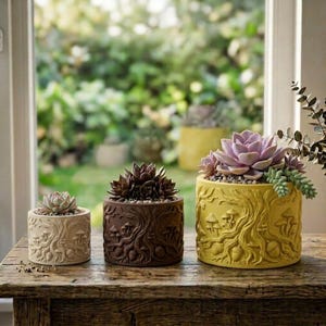 May include: Three cylindrical planters in cream, dark brown, and yellow, each featuring a relief design of tree roots, mushrooms, and acorns. The planters hold succulent plants of varying sizes, arranged in a row on a rustic wooden surface.