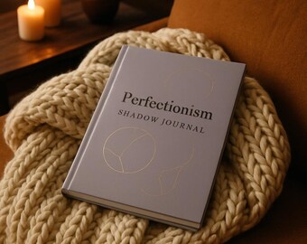 May include: A light gray hardcover journal with the title Perfectionism SHADOW JOURNAL printed in black text on the front cover, accompanied by thin gold line art illustrations of circular shapes resting on a thick cream-colored knit fabric.