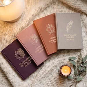 May include: Four hardback journals in purple, dusty rose, terracotta, and light grey, each featuring gold foil text and line art. The journals are titled Trauma-Aware Shadow Work Journal, Unmasked Shadow Work Journal, Nervous System Workbook, and Empath & HSP Shadow Journal.