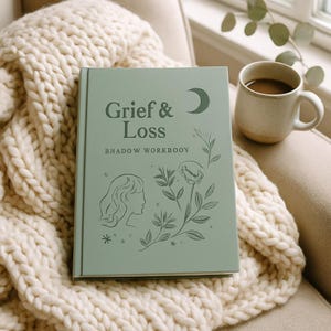 Könnte beinhalten: Ein salbeigrünes Hardcover-Arbeitsbuch mit dem Titel Grief & Loss BHADOW WORKBOOY, das auf der Vorderseite eine dunkelgrüne Strichzeichnung eines Profils, einer Blume und eines Halbmonds zeigt und auf einer dicken cremefarbenen Strickdecke liegt.