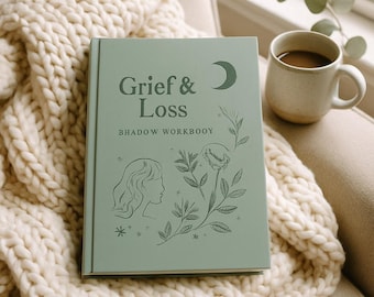 May include: A sage green hardcover workbook titled Grief & Loss BHADOW WORKBOOY, featuring a dark green line art illustration of a profile, a flower, and a crescent moon on the front cover, resting on a thick cream-colored knit blanket.
