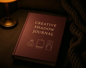 May include: A dark purple hardcover journal with the gold-colored text CREATIVE SHADOW JOURNAL printed on the front cover. Below the text are three simple gold-colored line drawings depicting an ink bottle, an easel, and a window with curtains.
