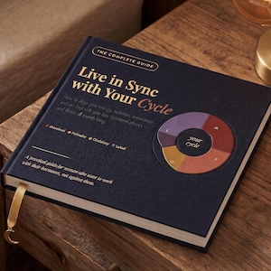 May include: A hardcover book titled "Live in Sync with Your Cycle" on a wooden surface. The cover is navy blue with gold lettering and a circular diagram. A gold ribbon bookmark with a crescent moon charm is visible.