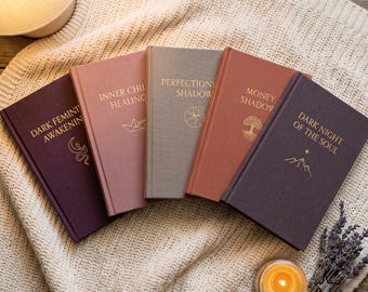 May include: Five hardcover books with gold foil lettering and symbols on their covers. The titles are Dark Feminine Awakening, Inner Child Healing, Perfectionism Shadow, Money Shadow, and Dark Night of the Soul, arranged in a fan shape on fabric.