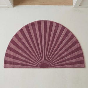 May include: A semi-circular rug featuring a radiating sunburst pattern in alternating shades of deep plum and dusty rose. The design consists of textured, raised stripes that fan out from the center of the flat edge toward the curved border.