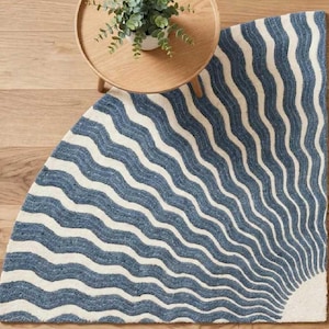Op de afbeelding: Een halfrond vloerkleed met een patroon van golvende blauwe en gebroken witte strepen die vanuit één hoekpunt uitstralen. Het kleed ligt onder een kleine, ronde houten tafel met daarop een groene plant in een pot.