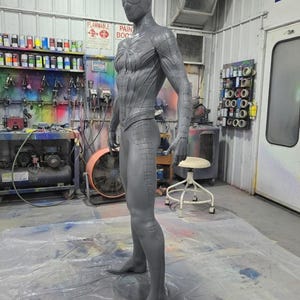 May include: A gray, full-body statue of a superhero in a form-fitting suit. The statue is in a workshop setting, with paint supplies and equipment visible in the background. The suit has a textured design.