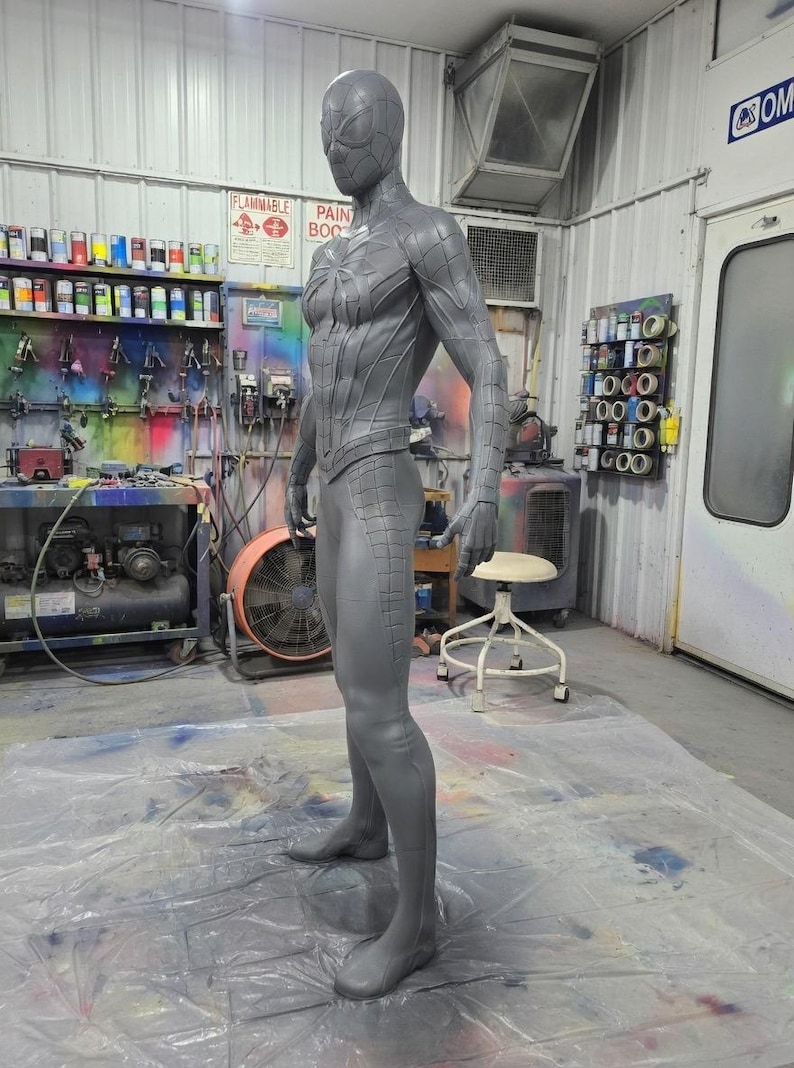 May include: A gray, full-body statue of a superhero in a form-fitting suit. The statue is in a workshop setting, with paint supplies and equipment visible in the background. The suit has a textured design.