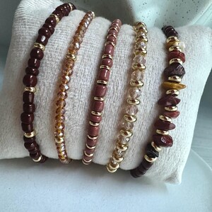 Peut inclure: Un ensemble de cinq bracelets extensibles composés de perles variées, incluant des perles rondes marron foncé, des cristaux ambrés facettés, des perles cylindriques vieux rose, des cristaux transparents facettés et des éclats de pierre rouge et jaune.