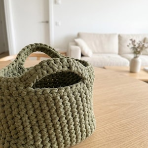 Peut inclure: Un petit panier en crochet vert olive avec deux anses. Le panier est fait de fil épais et texturé et repose sur une surface en bois clair. L'intérieur du panier est visible, et l'arrière-plan montre un cadre de salon.