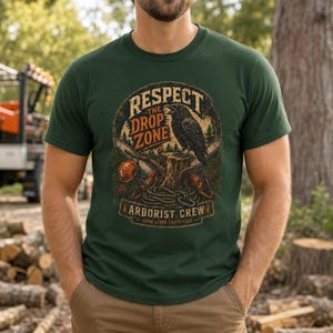 May include: A dark green t-shirt featuring a graphic print of a raven perched on a tree stump, chainsaws, and a rope. The text reads: RESPECT THE DROP ZONE ARBORIST CREW HIGH LIMB CERTIFIED. The design includes pine cones and forest elements.