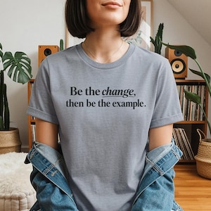 以下が含まれることがあります： 「Be the change, then be the example.」という黒い文字がプリントされた、ヘザーグレーの半袖Tシャツ。クルーネック仕様で、肩には青いデニムジャケットが羽織られており、袖と襟部分が見えています。