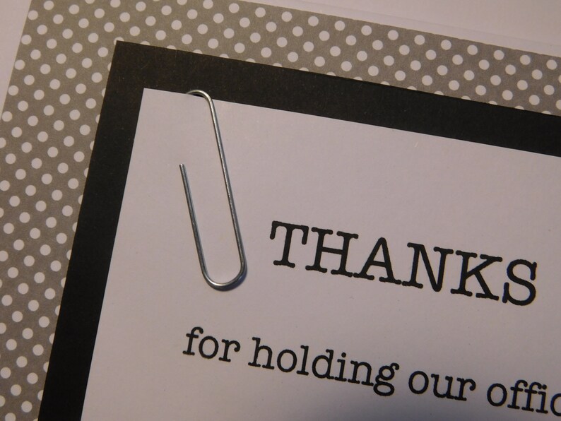 May include: A white card with black text that reads "THANKS for holding our offic", a silver paper clip is attached to the top of the card.
