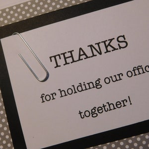 May include: A white card with black text and a silver paperclip. The text reads "THANKS for holding our office together!"