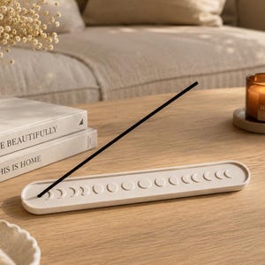 May include: A long, off-white, rectangular incense holder featuring a series of embossed moon phases along its centre. A single thin black incense stick is inserted into the holder at an angle, resting against the surface of the tray.