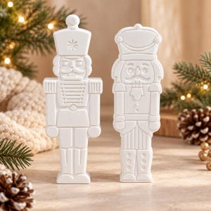 May include: Two matte white nutcracker figurines standing side by side. The left nutcracker features a tall hat with a star detail and a uniform with epaulets. The right nutcracker has a rounded hat and a detailed jacket with vertical stripes.