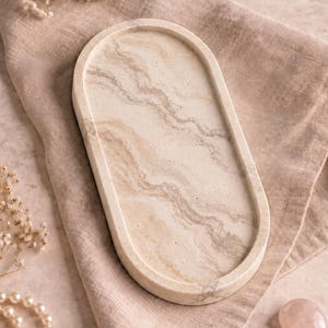 May include: An oval-shaped tray featuring a smooth, light beige surface with swirling cream and tan marble-like patterns. The tray has a raised, rounded edge and a solid, flat base, presenting a consistent colour palette throughout its structure.