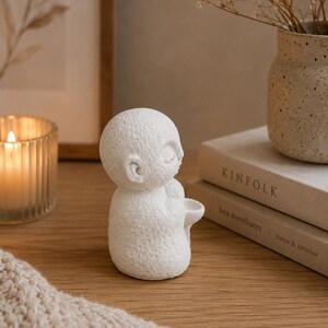 May include: A small, white, textured figurine of a seated figure with closed eyes, holding a small bowl in its hands. The sculpture has a matte, porous surface and a rounded, stylized head, positioned upright on a flat surface.
