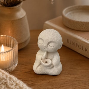 May include: A small, off-white, textured figurine of a seated figure with a smooth, rounded head and closed eyes. The figure is depicted in a meditative pose, holding a small, circular bowl-shaped object with a central hole in its hands.